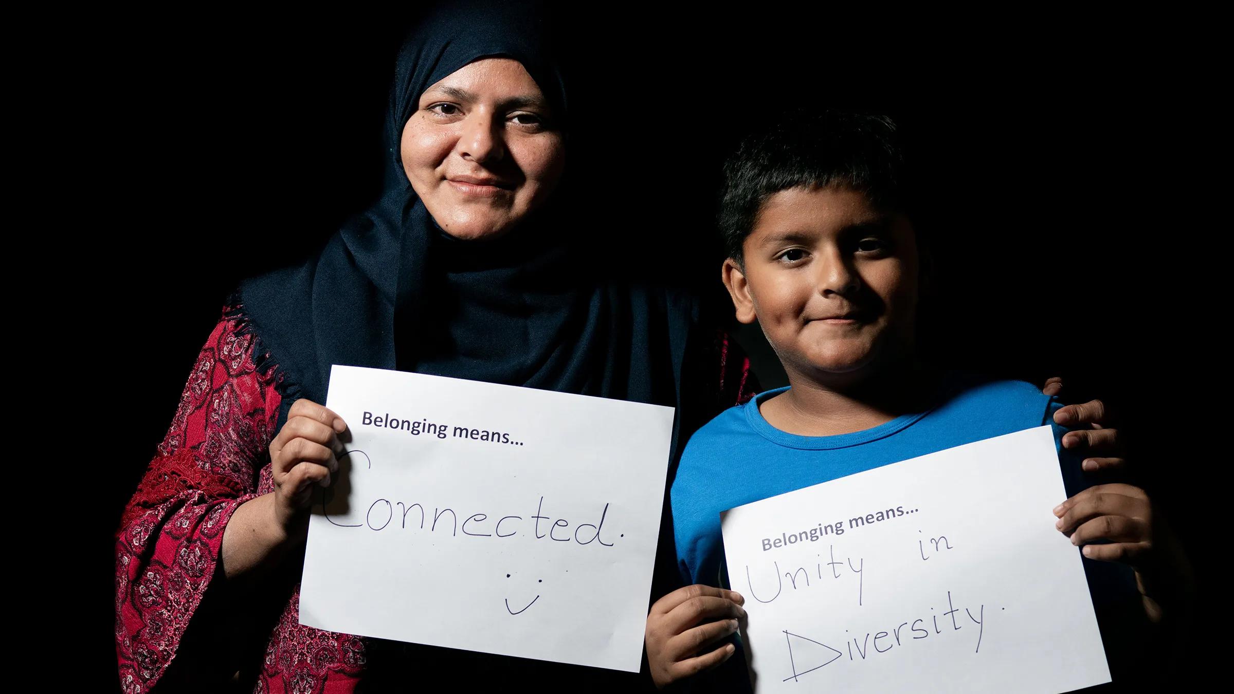 Mother and son smiling and with arms around each other, holding sings that say "Belonging is.." with their answers to the question - Connected and Unity in diversity.